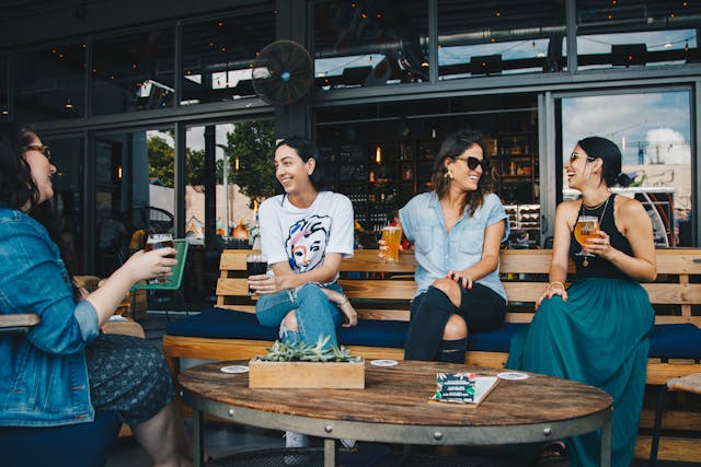 four-women-sitting-outside-at-a-wooden-table-smiling-and-chatting-while-holding-drinks-at-a-cafe